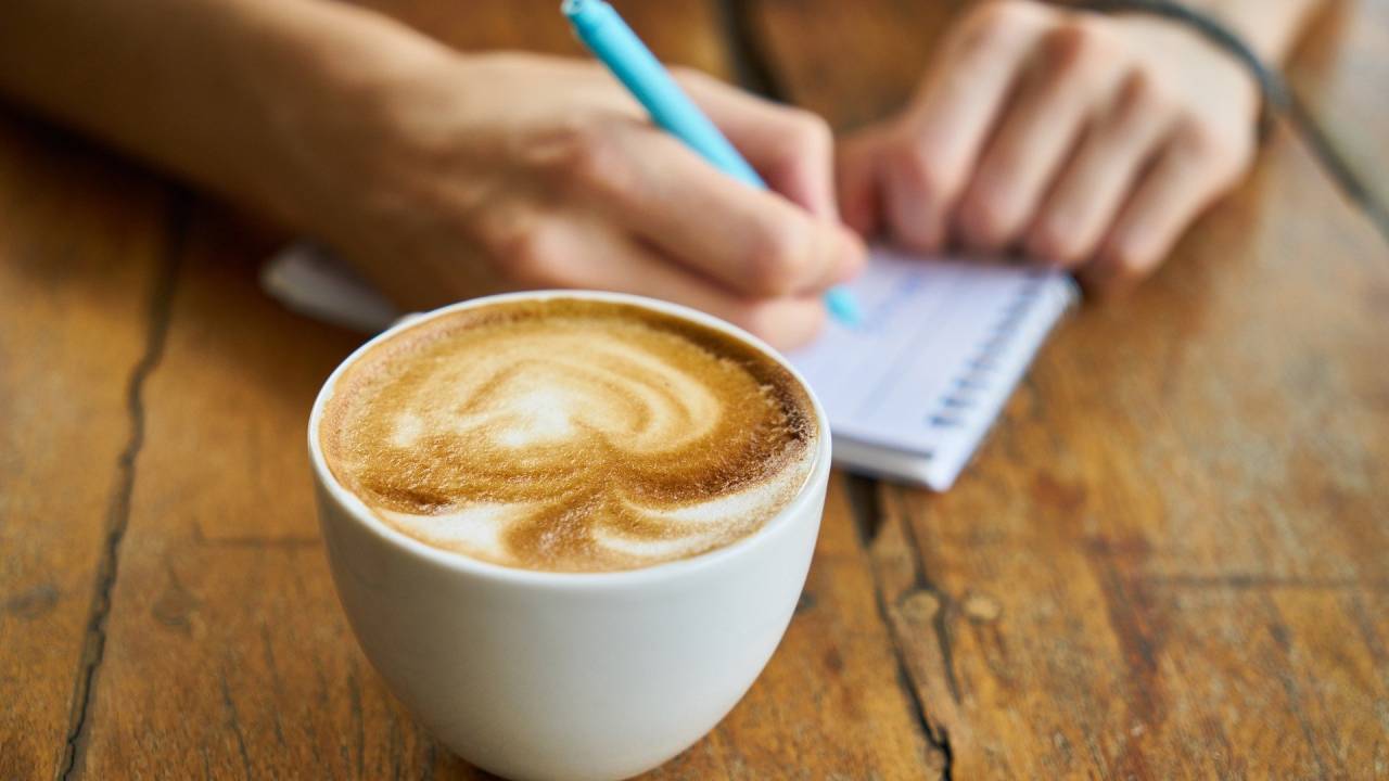 pessoa escrevendo em um bloco de notas com um café ao seu lado