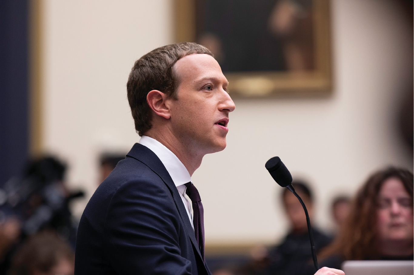 Zuckerberg, de perfil. Ele veste terno e está sentado em frente a um microfone.