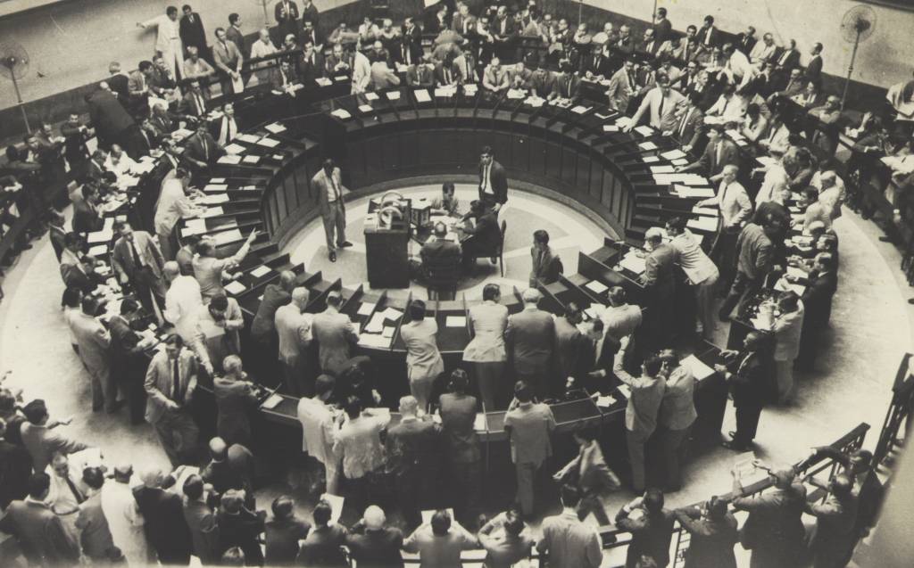 O pregão da Bovespa, atual B3, em foto de meados de século 20. Trata-se de uma grande mesa redonda, com vários homens de terno apoiados trocando papéis. A foto integra o acervo do Museu Paulista.