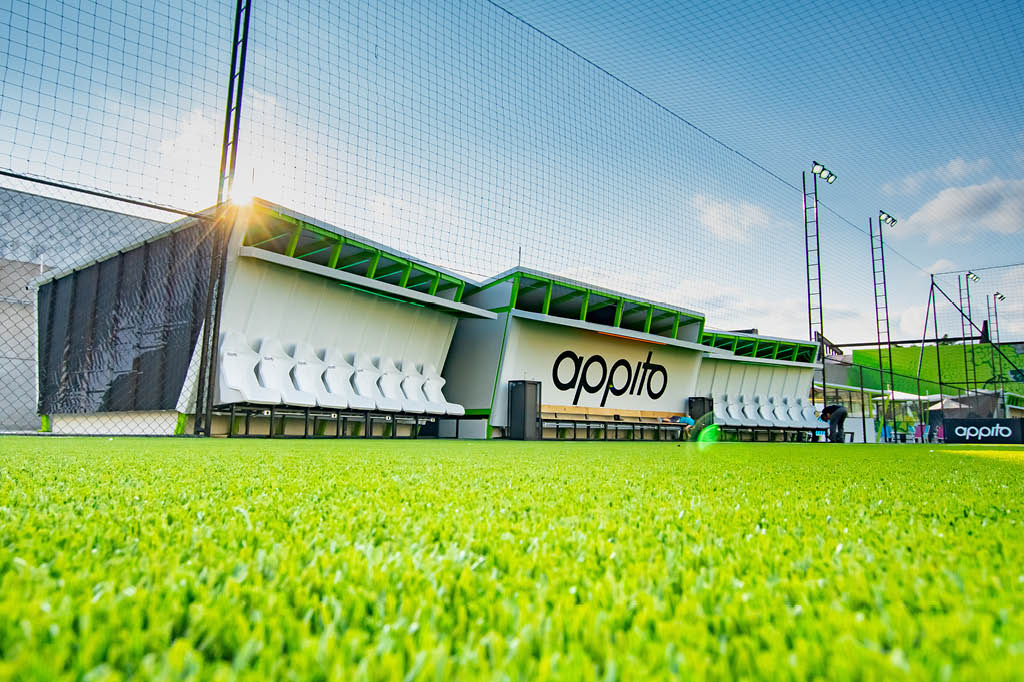 Fotografia de um campo de futebol da arena da Appito, mostrando o gramado e o espaço dos bancos de reservas.