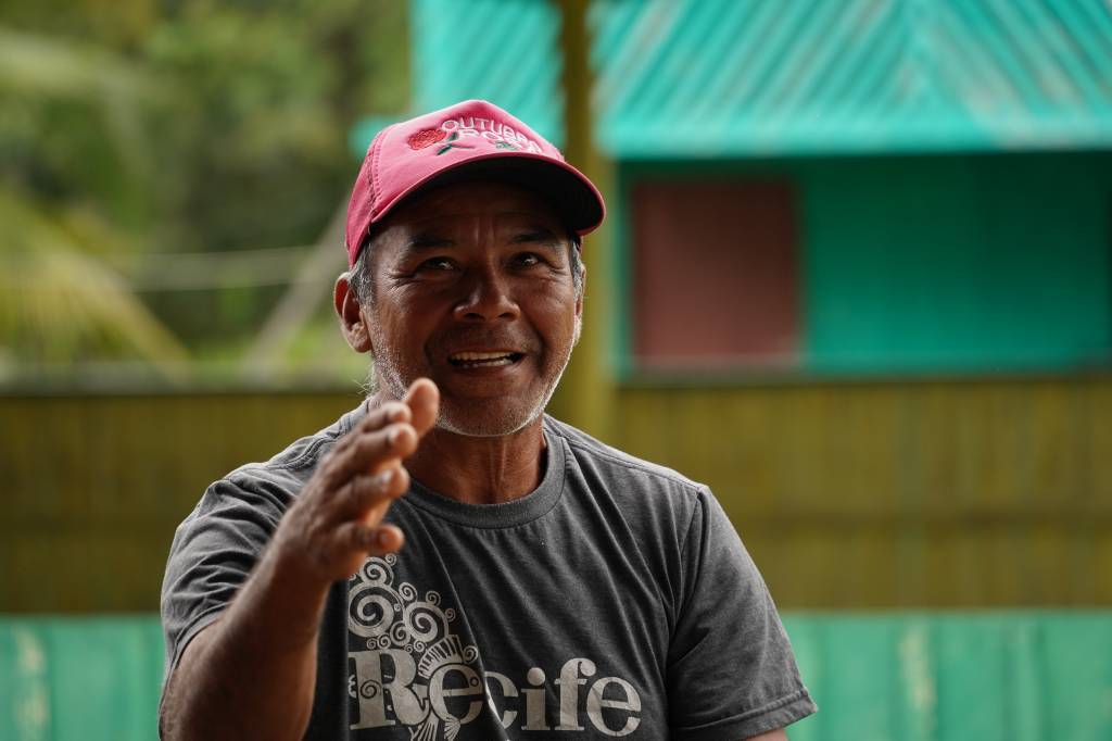Homem indígena de boné vermelho e camiseta verde, sentado sorrindo posando para a foto. Fundo desfocado