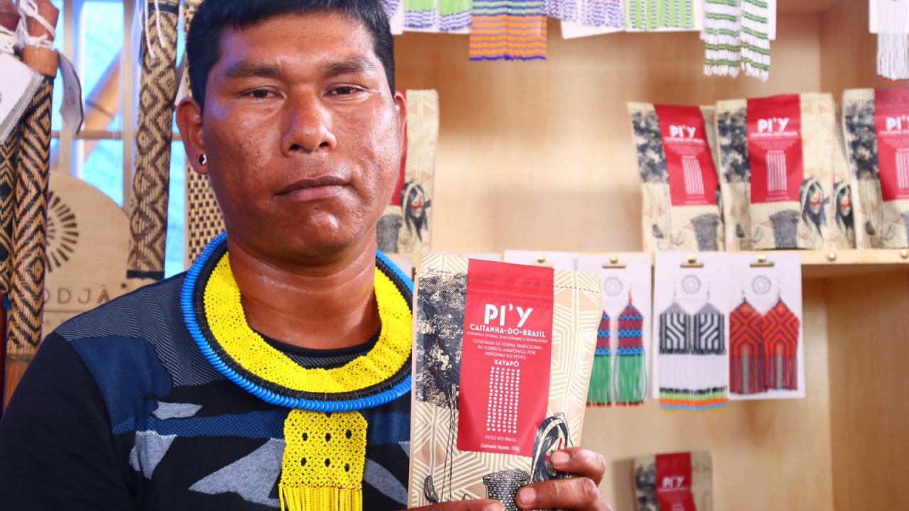 Homem indígena olha para a câmera e sorri levemente. Em suas mãos, um pacote bege e vermelho de castanha, comercializado pela plataforma.
