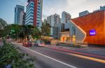 A imagem mostra uma avenida movimentada em uma área urbana com vários prédios altos ao fundo. À direita, há um edifício com fachada laranja e cinza, exibindo o logotipo do banco Itaú iluminado. A calçada à frente do prédio é decorada com vegetação, e carros passam pela rua, deixando rastros de luzes devido ao movimento rápido. O céu está claro, indicando que a foto foi tirada ao entardecer.