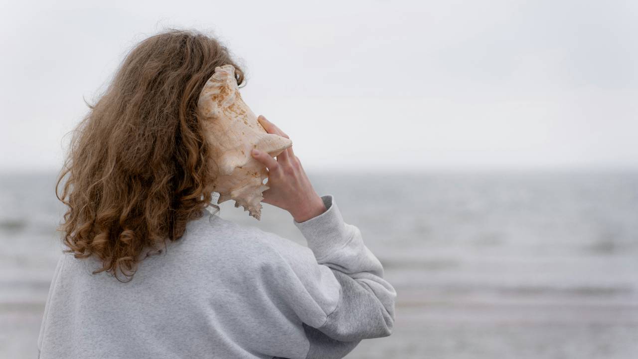A imagem mostra uma pessoa de costas, com cabelo ondulado e usando um moletom cinza. Ela está segurando uma concha grande junto à orelha, enquanto olha para o mar ao fundo. O cenário é tranquilo, com o céu nublado e o oceano levemente desfocado ao longe.