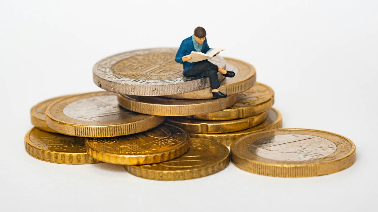 A imagem mostra uma figura em miniatura de uma pessoa sentada em cima de uma pilha de moedas douradas, lendo um jornal. A figura está vestida com roupas formais, e a cena é capturada em um fundo branco, destacando o contraste entre o pequeno tamanho da figura e as moedas maiores.