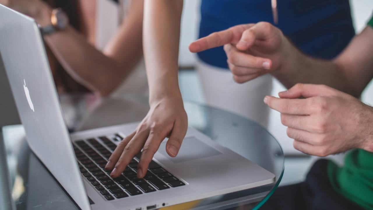 A imagem mostra três pessoas reunidas ao redor de um laptop, com foco em suas mãos e braços. Uma pessoa está digitando no teclado do laptop, enquanto outra está apontando para a tela, como se estivesse explicando ou discutindo algo. O laptop está sobre uma mesa de vidro, e as mãos de todas as pessoas na imagem estão próximas ao dispositivo. A imagem sugere um ambiente de colaboração ou trabalho em equipe.