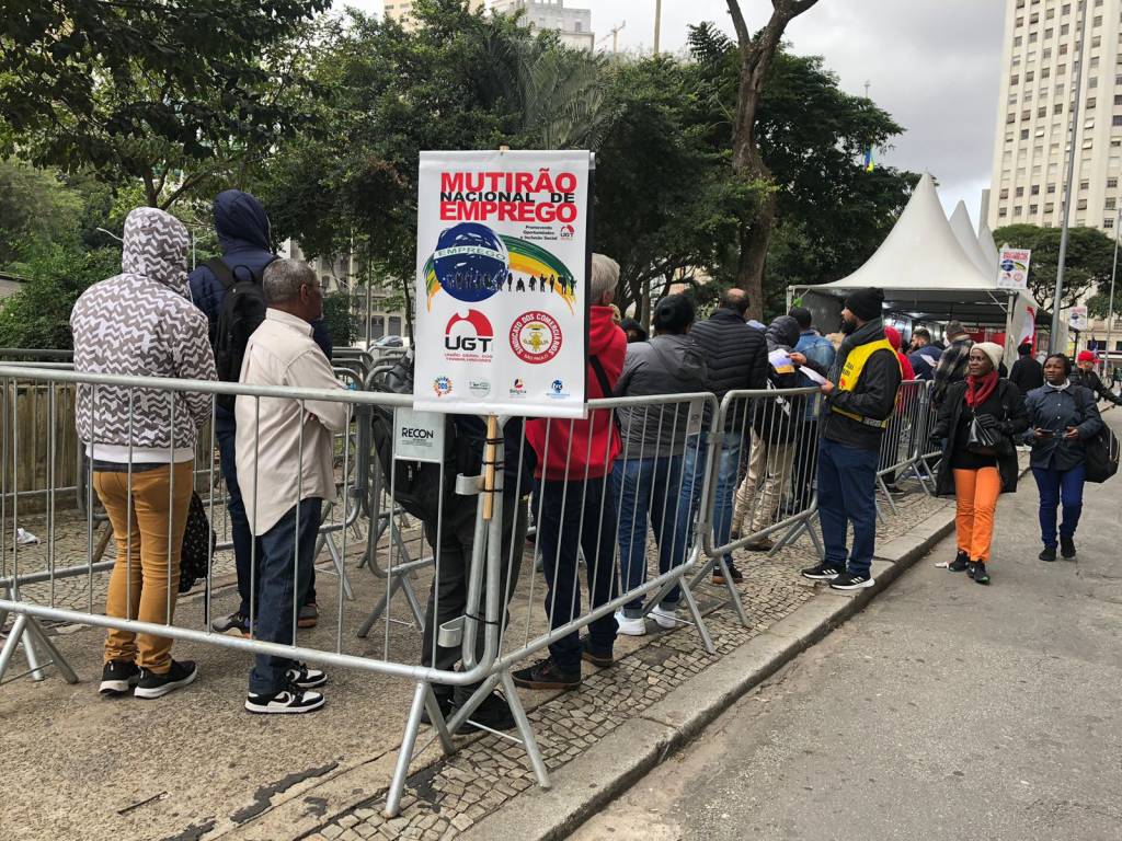 A imagem mostra uma fila de pessoas ao ar livre, aguardando para participar de um mutirão nacional de emprego. As pessoas estão vestidas com roupas de frio, indicando que o clima é ameno ou frio. Algumas delas estão encostadas em grades de contenção, que organizam a fila. No centro da imagem, há um cartaz com a inscrição 