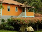 casa amarela na frente de uma floresta com várias arvores altas e verdes. embaixo da casa, um toldo rodeado por flores rosas claras, e um fusca amarelo estacionado embaixo.