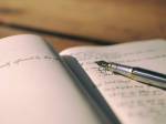 A imagem mostra um caderno aberto com uma caneta-tinteiro prateada apoiada sobre as páginas. O caderno tem linhas e contém anotações escritas à mão. A cena está desfocada ao fundo.