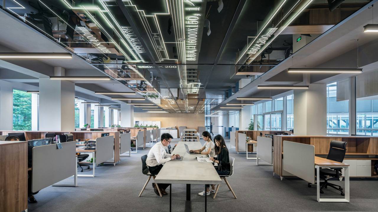 A imagem mostra um escritório moderno e espaçoso com várias baias de trabalho. No centro, três pessoas estão sentadas em uma mesa compartilhada, usando laptops. O teto tem um design reflexivo com linhas e textos iluminados. As estações de trabalho nas laterais possuem divisórias baixas, e há janelas grandes ao fundo que permitem a entrada de luz natural. O ambiente é organizado, com iluminação artificial suave e plantas decorativas em algumas mesas.