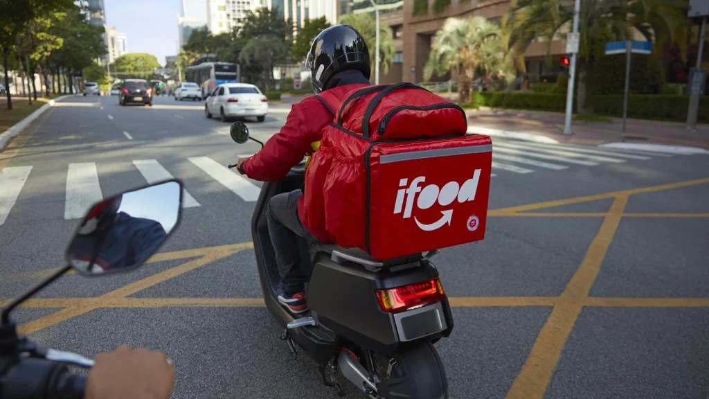 A imagem mostra um entregador de motocicleta usando uma mochila vermelha do iFood, um serviço de entrega de alimentos. Ele está pilotando pela rua de uma cidade, usando capacete preto e jaqueta vermelha. À frente dele, há outros veículos na estrada, e o cenário urbano inclui árvores, prédios e sinalização de trânsito.