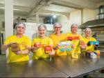 A imagem mostra cinco pessoas em uma cozinha industrial, todas vestindo camisetas amarelas e toucas de proteção nos cabelos. Elas estão sorrindo e seguram embalagens de comida prontas para entrega, exibindo-as para a câmera. Atrás delas, é possível ver alguns equipamentos de cozinha, como fornos e prateleiras.