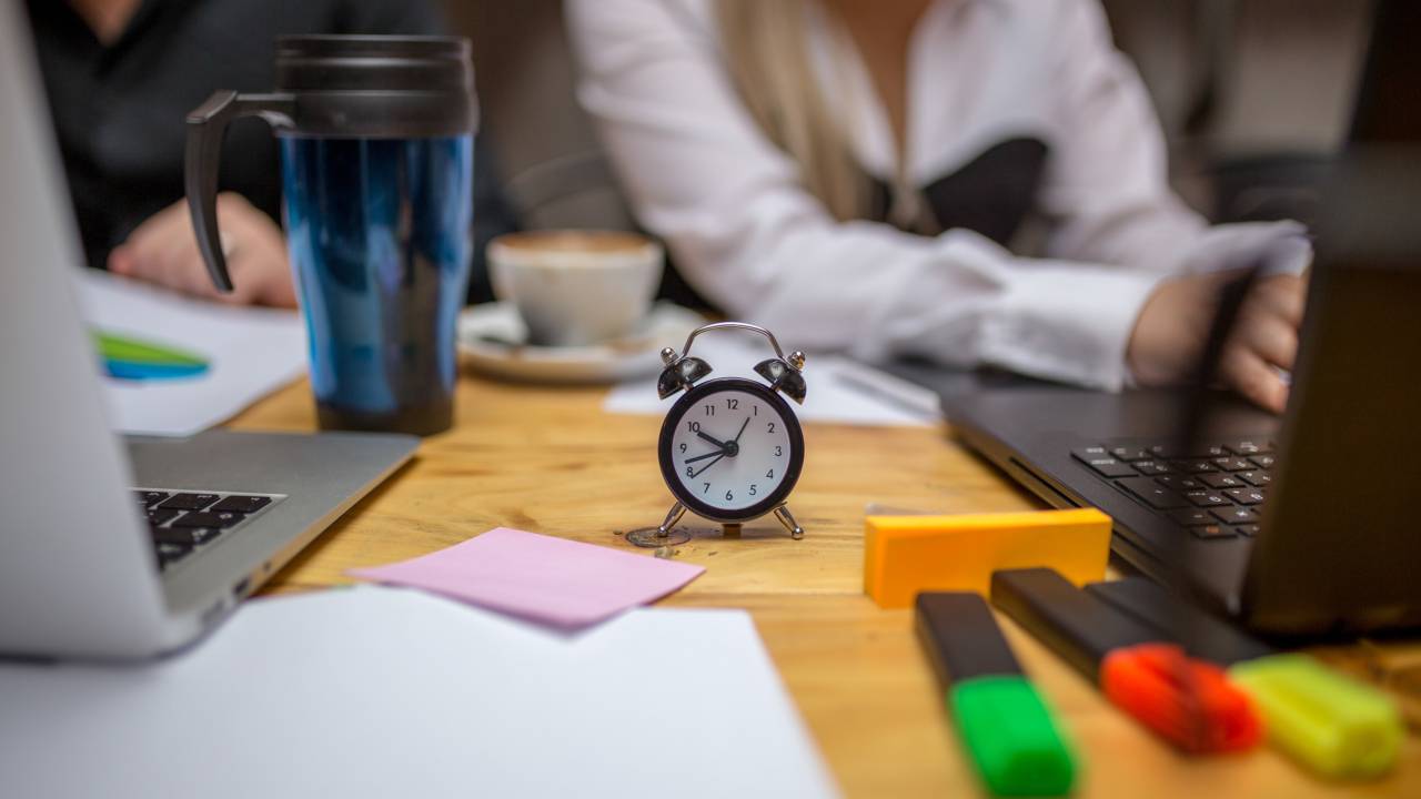 Empresários trabalhando em uma mesa de trabalho cheio de materiais, a imagem foca em relógio analógico que está no centro da mesa.