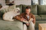 Fotografia de um homem lendo um livro em sua sala de estar, acompanhado de seu cachorro.