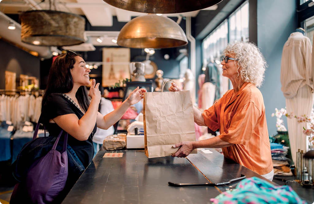 Fotografia de um mulher entregando sacola a cliente no caixa de uma loja de roupas.