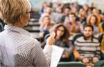 Uma professora lecionando uma aula. Elas está de costas, com uma mão estendida e a outra, segurando uma folha de papel. Ela veste óculos e possui cabelos curtos.