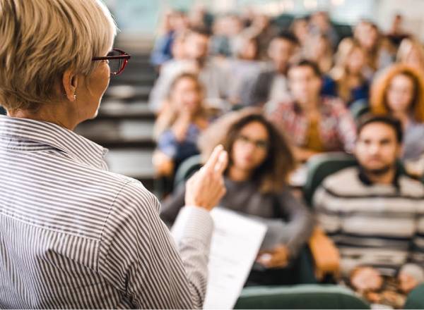 Uma professora lecionando uma aula. Elas está de costas, com uma mão estendida e a outra, segurando uma folha de papel. Ela veste óculos e possui cabelos curtos.