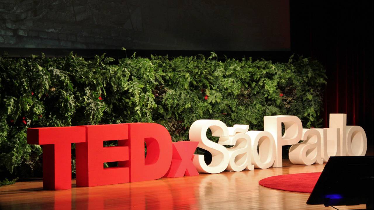 Fachada com letreiro escrito "TEDxSão Paulo", nas cores vermelha e branca, posicionado na frente de uma parede com folhas e plantas.