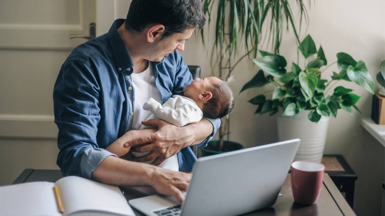 Homem segurando bebê enquanto digita algo em notebook.