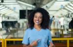 Uma mulher negra de cabelos cacheados vestindo uniforme azul com o logo da Volkswagen. Ela está apontando para o logo com os seus dados indicadores.